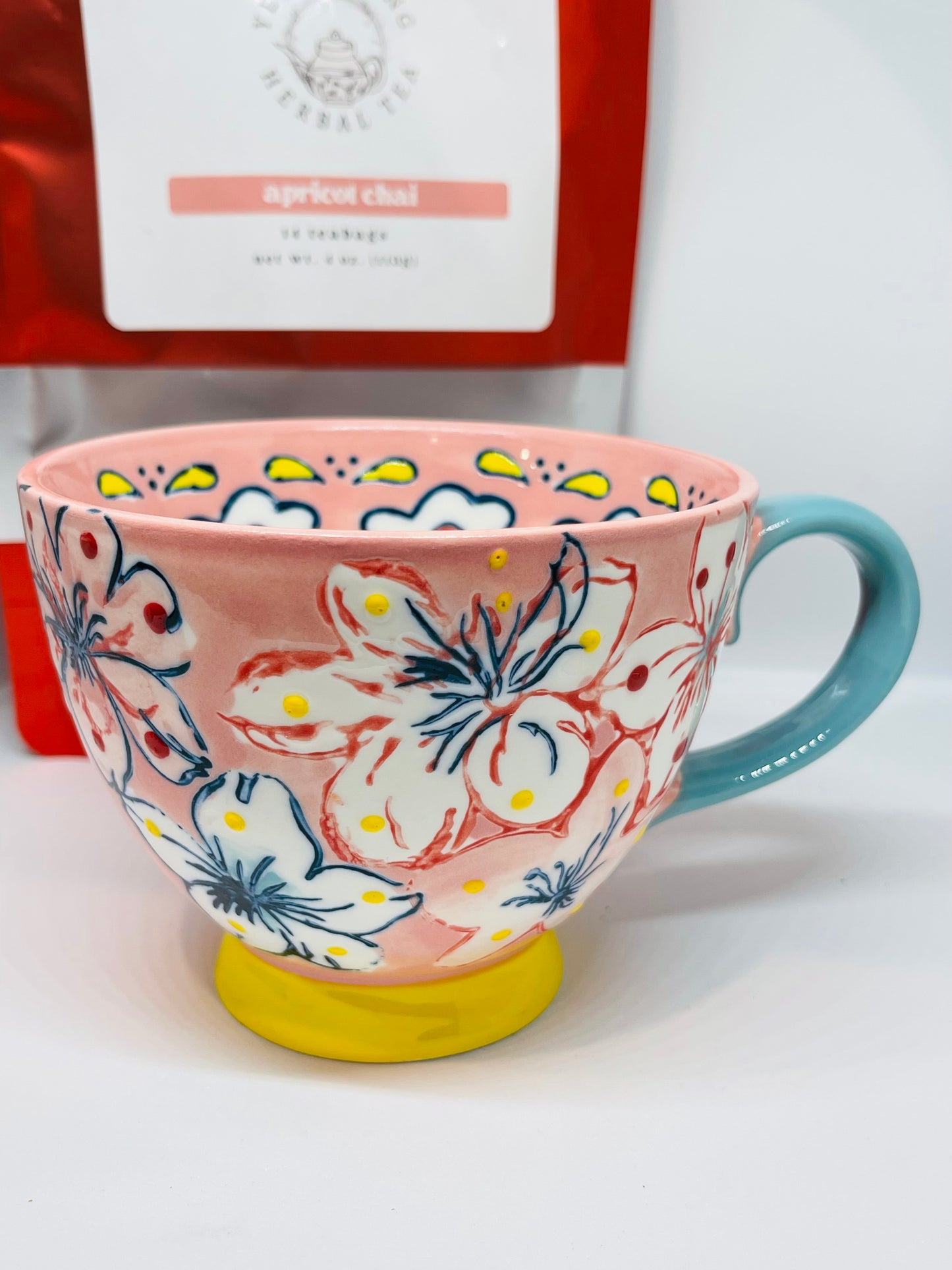 A light pink tea mug with red and navy blue trimmed white and pink flowers with yellow and red dots, a yellow base, and a turquoise handle.