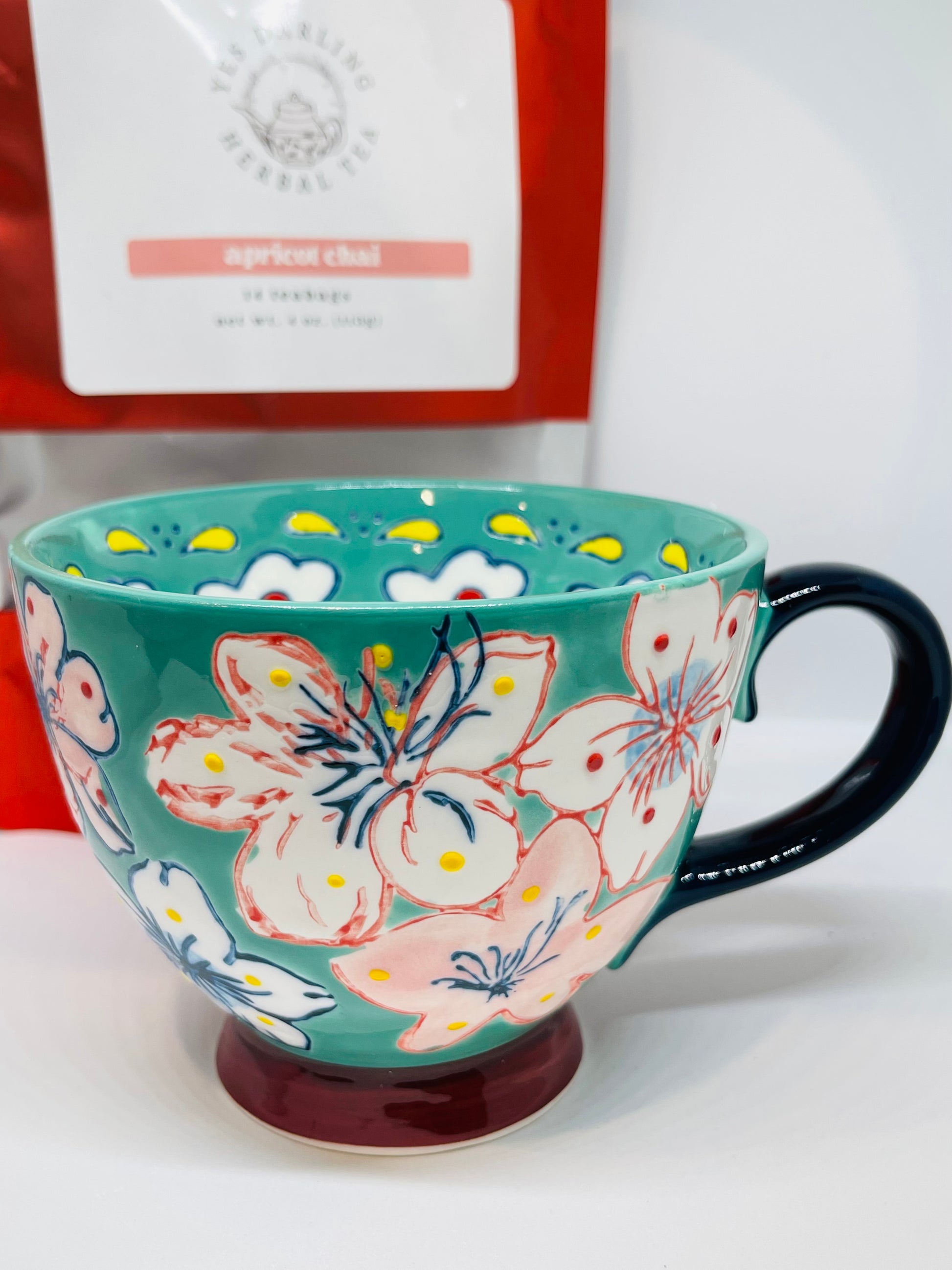 A teal mug with red and navy blue trimmed white, pink, and blue flowers with red and yellow dots, a navy blue handle, and a burgundy base.