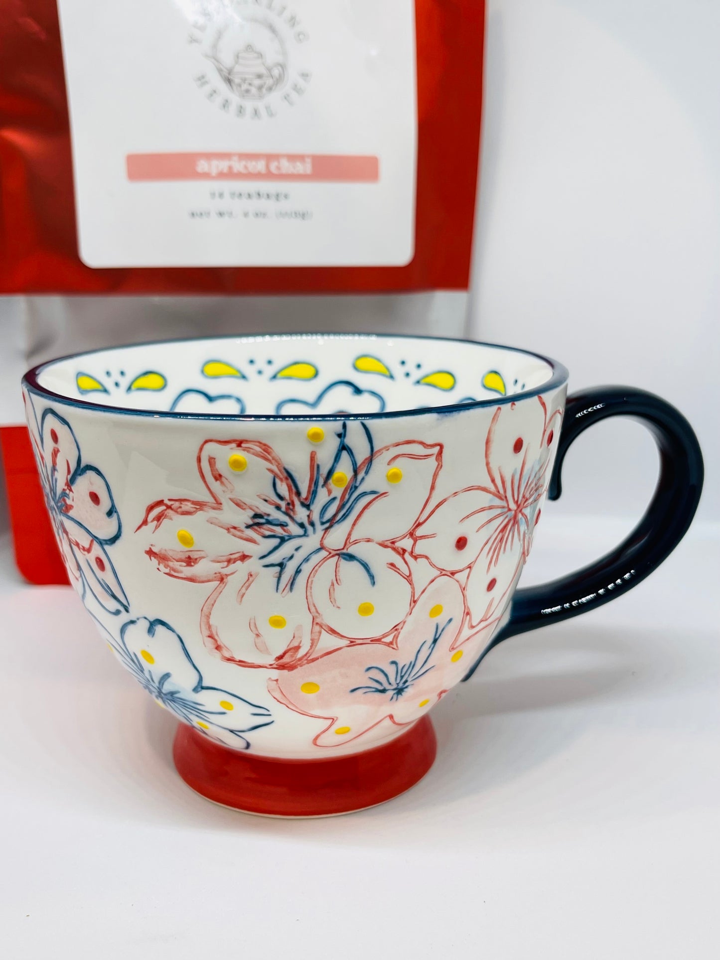A white mug with red and navy blue trimmed flowers with yellow and red dots, a navy blue rim, a red handle, and a navy blue base.