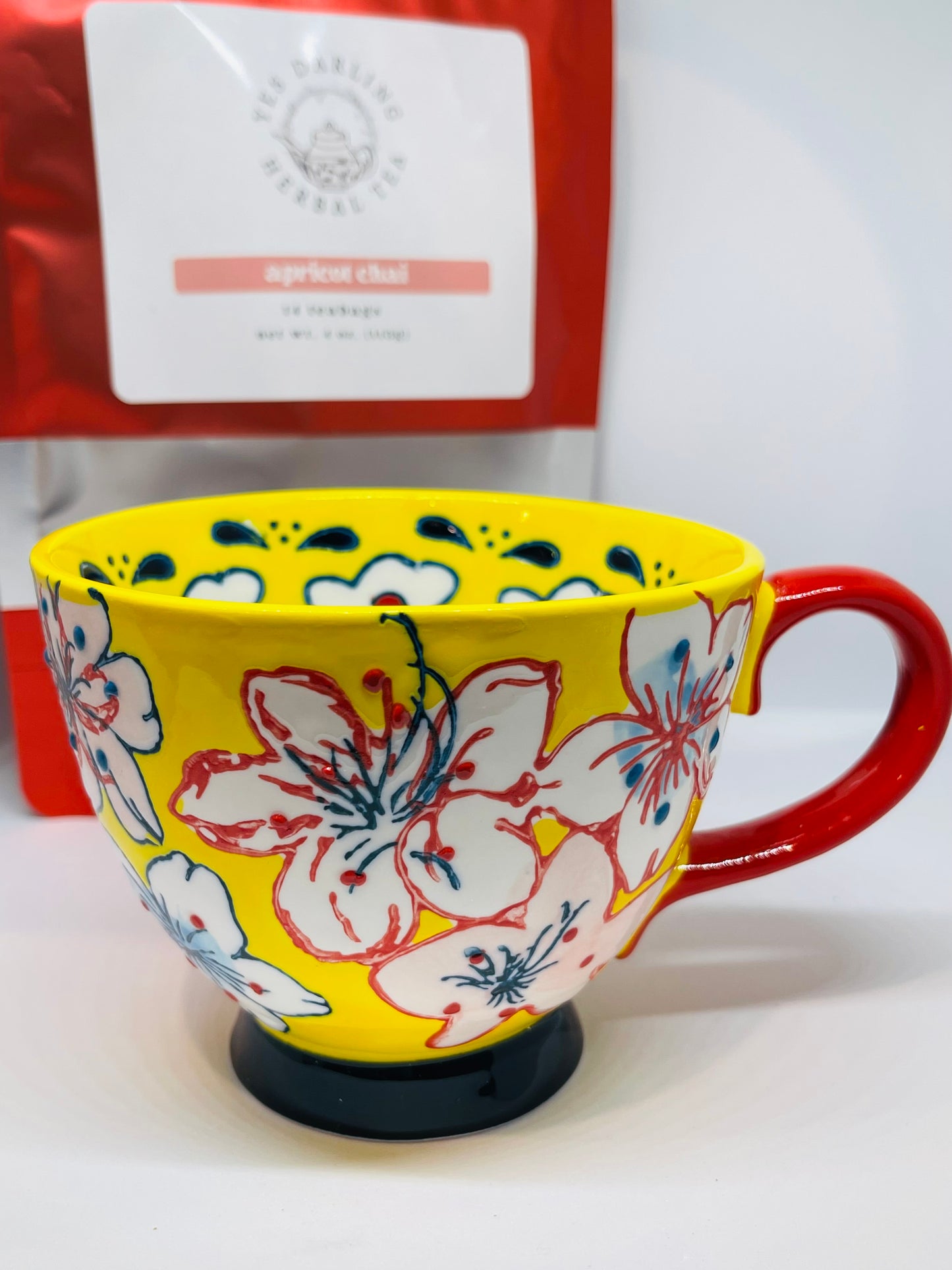 A yellow tea mug with red and navy blue trimmed white flowers with pink and blue spots and red and navy blue dots, a red handle, and a navy blue base.